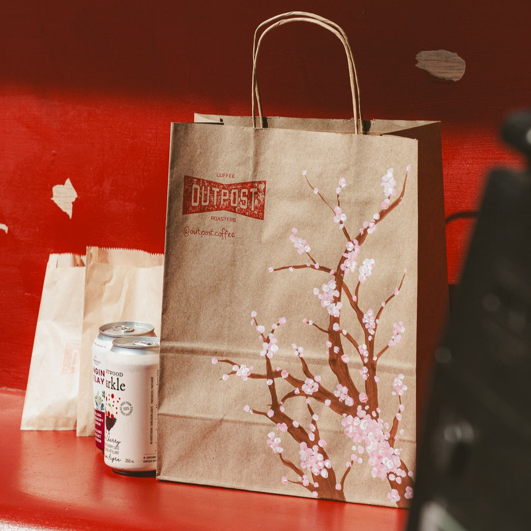 A paper picnic lunch bag with a cherry blossom design, accompanied by a smaller paper bag and a can, displayed on a red bench