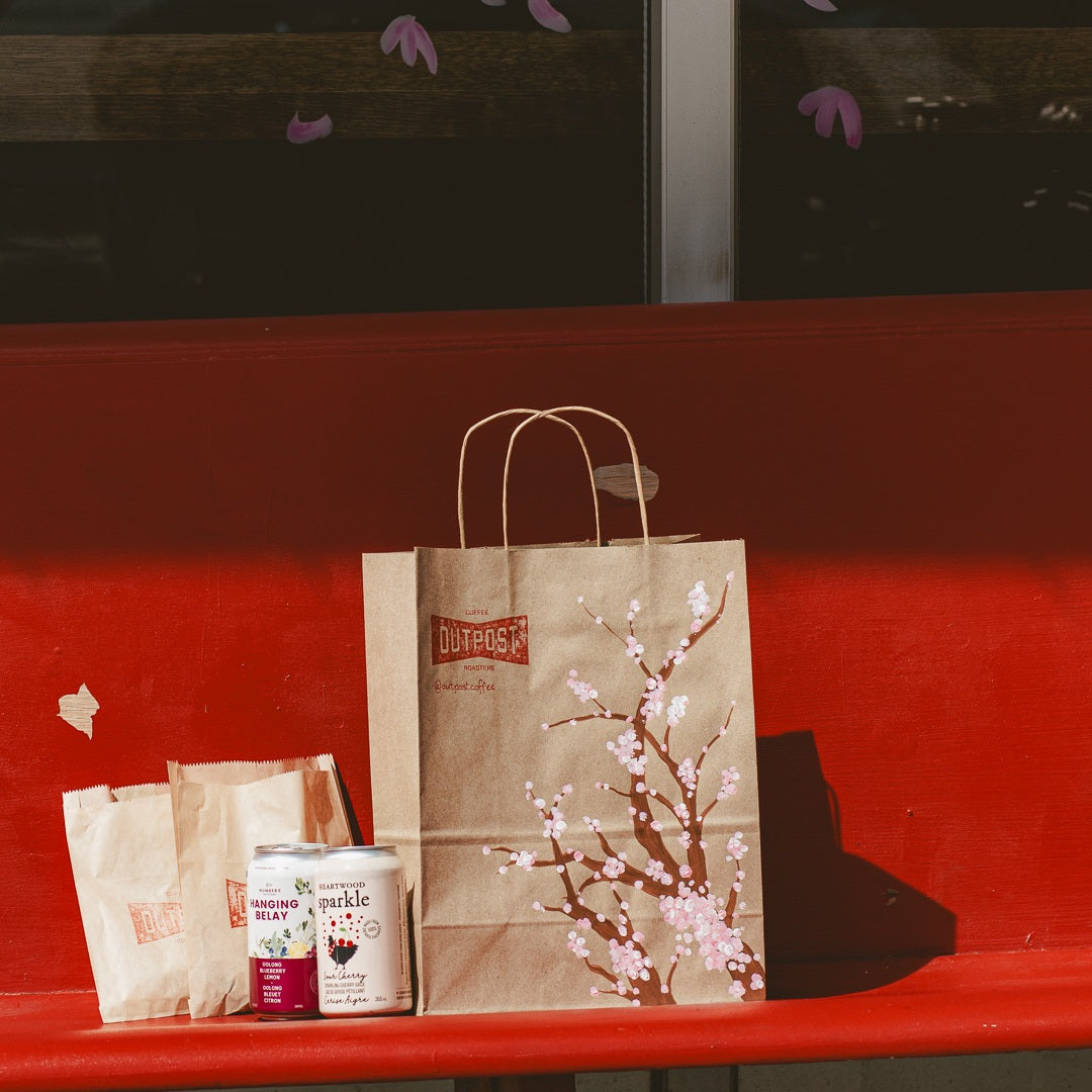 A paper picnic lunch bag with a cherry blossom design, accompanied by a smaller paper bag and a can, displayed on a red bench with a window in the background.