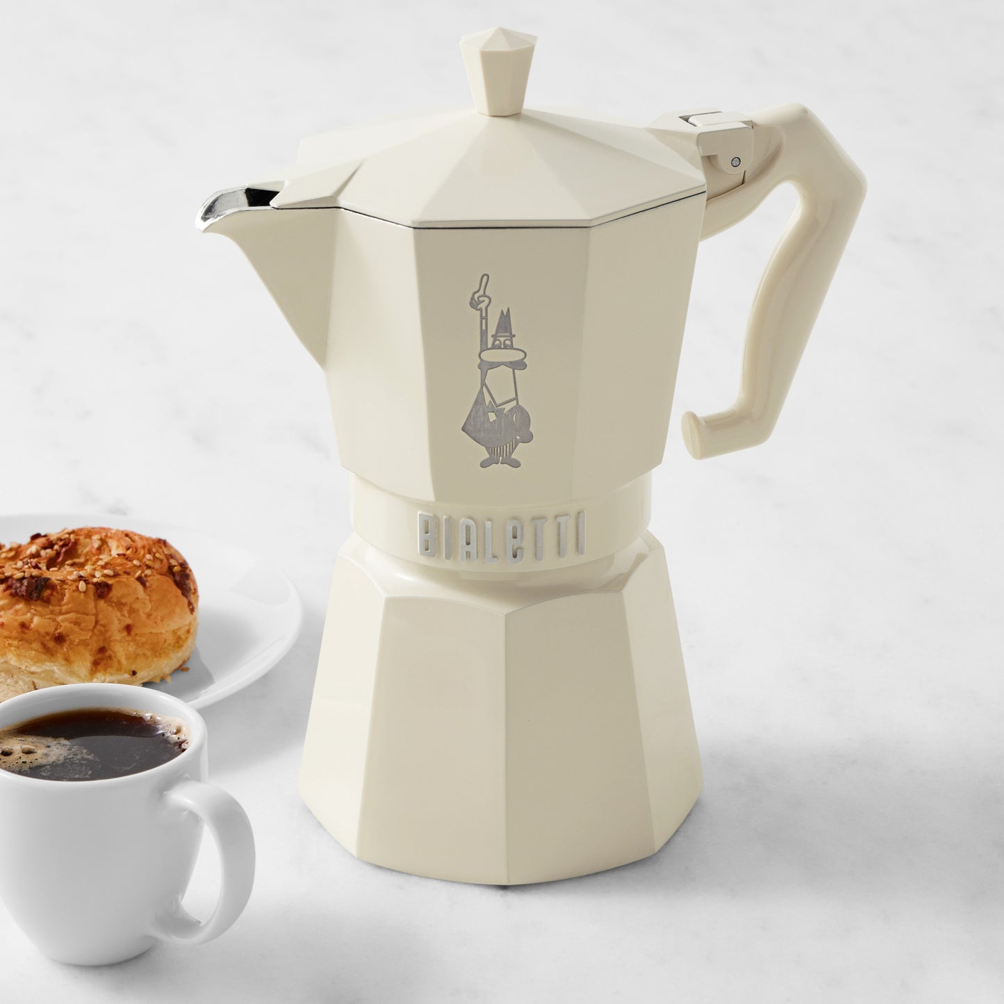 Cream coloured Bialetti coffee maker with a cup of coffee and pastry on a white background