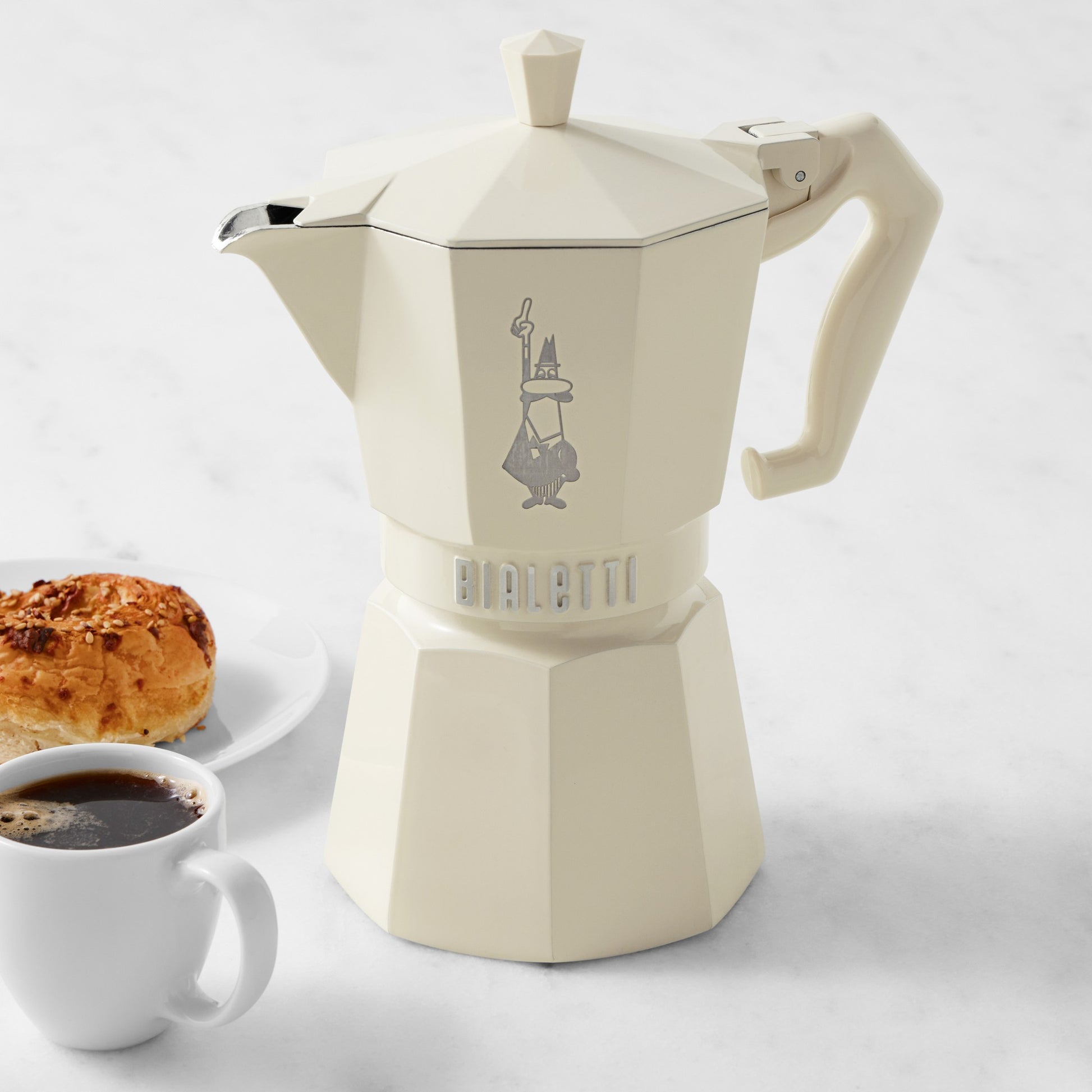 Cream coloured Bialetti coffee maker with a cup of coffee and pastry on a white background